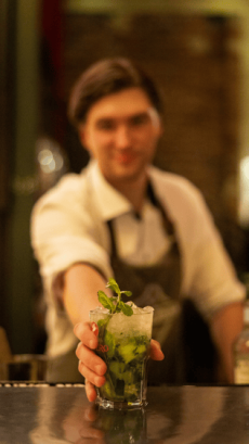 Bild von einer verschwommenen Person im Hintergrund, die einen Cocktail nach vorne ins Bild stellt.